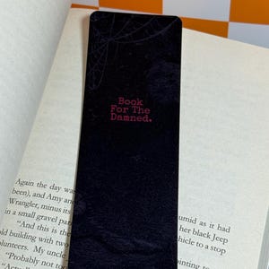 May include: A rectangular black bookmark featuring a subtle dark spiderweb pattern at the top and the red text "Book For The Damned." printed in the center. The bookmark is placed vertically against the open pages of a printed book.