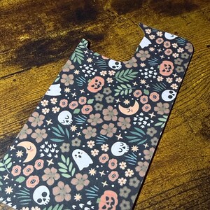 May include: A decorative phone case featuring a dark background with a repeating pattern of skulls, ghosts, pumpkins, flowers, stars, and crescent moons in shades of peach, white, and green. The case has a unique shape with cutouts for the camera and buttons.