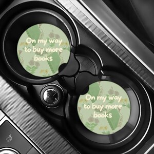 May include: Two car coasters with a light green background, floral pattern, and the text "On my way to buy more books." The coasters are round and fit into a car's cup holders. The car's interior is black and gray.
