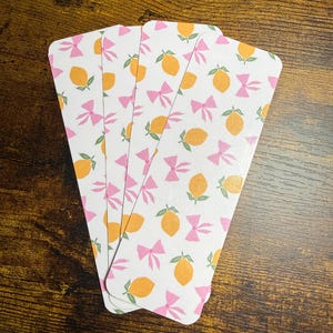 Peut inclure: Quatre marque-pages blancs aux coins arrondis, chacun avec un motif répété de citrons oranges, de nœuds roses et de feuilles vertes. Les marque-pages sont disposés en éventail sur un fond en bois.