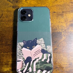 Peut inclure: Coque de téléphone transparente avec un dessus vert teal et un graphique d'un chat noir lisant un livre. Le livre porte le texte "Comment Domestiquer Les Humains". Le chat est allongé sur un oreiller et couvert d'une couverture.