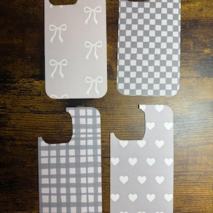 Pode incluir: Quatro capas de telefone com diferentes padrões. Uma tem laços brancos em um fundo cinza. Outra tem um padrão de xadrez cinza e branco. A terceira tem um design xadrez cinza e branco. A última tem corações brancos em um fundo cinza.