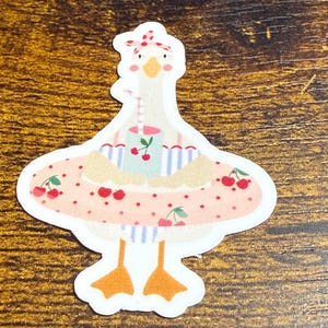 May include: A die-cut sticker featuring a cartoon goose wearing a pink inner tube with cherries. The goose is holding a drink with cherries and wearing a red and white polka dot bow. The goose has orange feet and a white body.