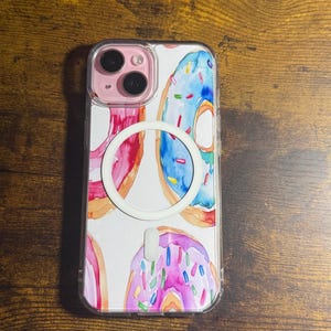 Puede incluir: Funda transparente para teléfono con un diseño de acuarela de donuts en tonos rosa, azul y naranja. La funda tiene un anillo MagSafe blanco y un módulo de cámara rosa.