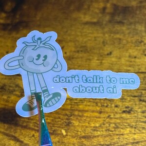 May include: A light blue sticker featuring a cartoon tomato character with the text "don't talk to me about ai". The tomato has a smiling face and is wearing shoes. The sticker is held by a pair of tweezers.