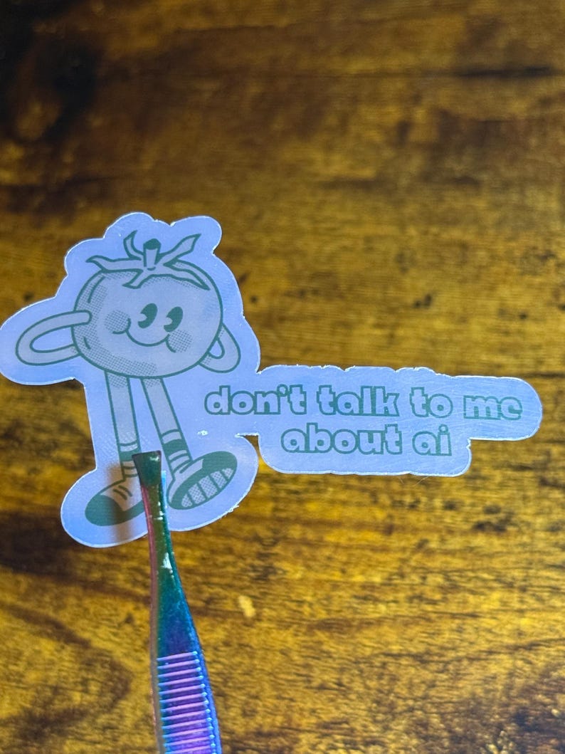 May include: A light blue sticker featuring a cartoon tomato character with the text "don't talk to me about ai". The tomato has a smiling face and is wearing shoes. The sticker is held by a pair of tweezers.