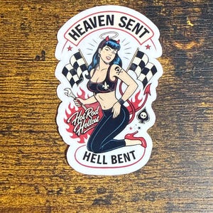May include: A sticker featuring a retro pin-up girl with devil horns and a halo, holding a wrench and posing with checkered flags. The text reads "Heaven Sent" and "Hell Bent" with "Hot Rod Hellcat" in a flame design.