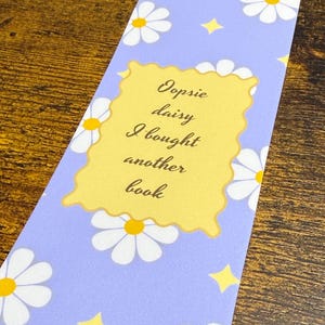 May include: A rectangular bookmark with a light purple background featuring a repeating pattern of white daisies with yellow centers and small yellow stars. A central yellow scalloped rectangle contains the black text: Oopsie daisy I bought another book.