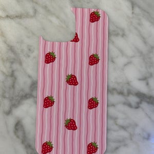 May include: A pink phone case with a vertical striped pattern and scattered red strawberries with green leaves. The case has a cutout for the camera and other phone features.