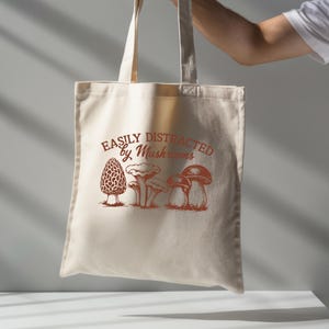 Puede incluir: Bolso tote beige con un gráfico marrón de varias setas y el texto "EASILY DISTRACTED by Mushrooms". El bolso está sostenido por una persona. El bolso está hecho de una tela de color claro.
