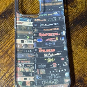May include: Clear phone case featuring a collage of VHS tapes with horror movie titles like "Halloween," "Friday the 13th," and "The Thing." The case has a transparent border and a design that covers the back.