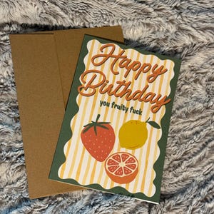 May include: A birthday card with a brown envelope. The card has a green border and a yellow and white striped background. The words "Happy Birthday you fruity fuck" are written in orange script. Illustrations of a strawberry, lemon, and orange are included.