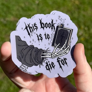 Op de afbeelding: Een lichtpaarse sticker met de tekst "This book is to die for" in een gotische lettertype. De sticker toont een zwart-wit illustratie van een skelet hand die een boek vasthoudt, met een zwarte mouw en arm.