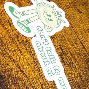 May include: A die-cut sticker featuring a cartoon character with a smiling face and limbs, wearing green shoes and striped socks. The sticker has the text "don't talk to me about ai" in green. The character is light green and white.