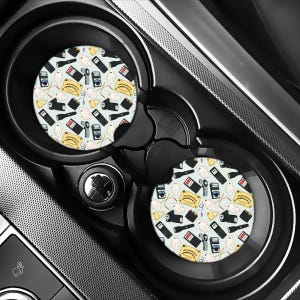 May include: Two circular car cup holder coasters featuring a light blue background with a repeating pattern of white ghosts, yellow ouija boards, black flashlights, and various ghost-hunting equipment including digital meters and sensors.