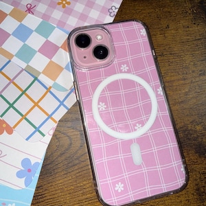 May include: A pink phone case with a plaid pattern and small white flower accents. The case has a clear border and a white circular MagSafe charger. The phone case is next to patterned paper.