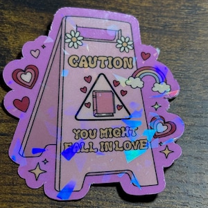 May include: A holographic sticker in the shape of a caution sign. The sign is lavender with the words "CAUTION" and "YOU MIGHT FALL IN LOVE". The sign has a book, hearts, daisies, a rainbow, and stars.