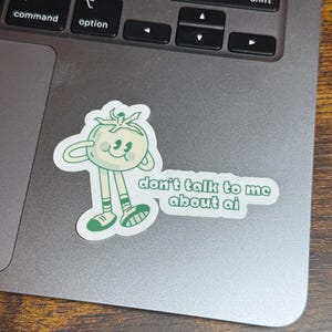 May include: A sticker featuring a cartoon tomato character with arms and legs. The tomato is light green with a smiling face and the text "don't talk to me about ai" in green letters.