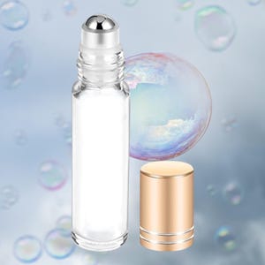 Puede incluir: Un frasco roll-on de vidrio transparente con una parte superior de bola rodante plateada y una tapa dorada separada. La botella est&aacute; vac&iacute;a, sobre un fondo degradado azul suave con burbujas flotantes.