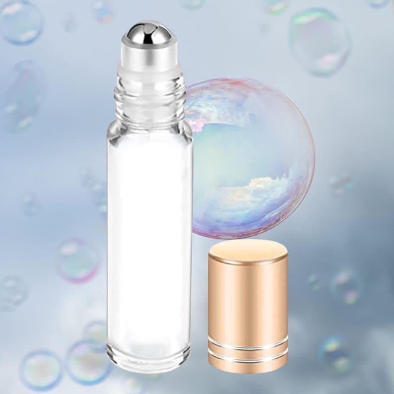 Puede incluir: Un frasco roll-on de vidrio transparente con una parte superior de bola rodante plateada y una tapa dorada separada. La botella est&aacute; vac&iacute;a, sobre un fondo degradado azul suave con burbujas flotantes.