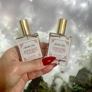 May include: Two clear glass perfume bottles with gold caps are held in a hand. The bottles are labeled "Apothic Luxe" with the text "BOOK SLUT" and "GOOD GIRL". Each bottle contains 30 ml of perfume.