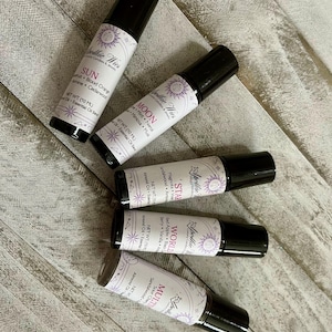 May include: Five black roller bottles with white labels are displayed on a light wood surface. Each label features the text "Apollie Wies" and a unique name: SUN, MOON, STAR, WORLD, and MULTI. The labels also list essential oil ingredients.