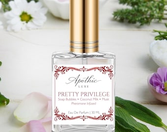 May include: A clear glass perfume bottle with a gold cap and a white label featuring red decorative borders. The label reads Apothic Luxe, Pretty Privilege, Soap Bubbles, Coconut Milk, Musk, Pheromone Infused, and Eau De Parfum | 30 ML.