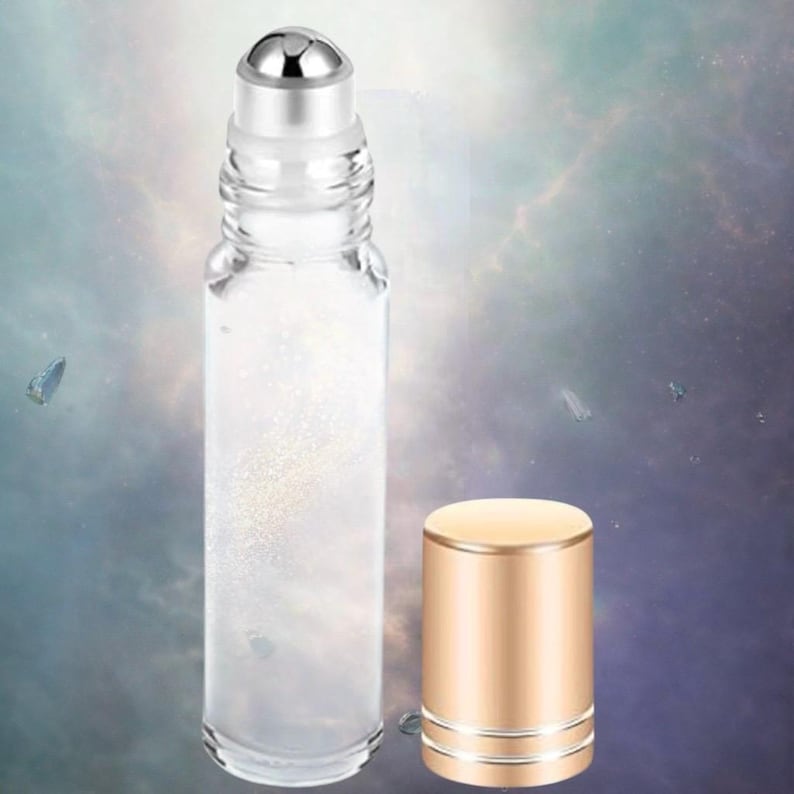 K&ouml;nnte beinhalten: Eine transparente Glas-Rollerflasche mit silbernem Rollballaufsatz und separater goldfarbener Kappe. Die Flasche enth&auml;lt eine schimmernde, glitzernde Fl&uuml;ssigkeit. Der Hintergrund ist ein weicher, verschwommener Farbverlauf aus Blau- und Lilat&ouml;nen.