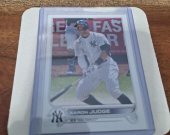 May include: A baseball card featuring Aaron Judge of the New York Yankees in a clear protective sleeve. The card shows Judge in his uniform, mid-swing. The Yankees logo is visible.