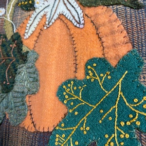 May include: A close-up of a handmade felt appliqué featuring a pumpkin, leaves, and flowers. The pumpkin is orange with a white stem and is surrounded by green leaves with gold stitching. The flowers are white with yellow centers.