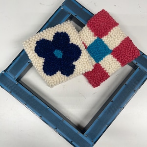 May include: Two small, square, hand-tufted textile pieces. One features a dark blue flower on a cream background, the other a red and cream checkerboard pattern with a turquoise square. A blue frame surrounds the pieces.