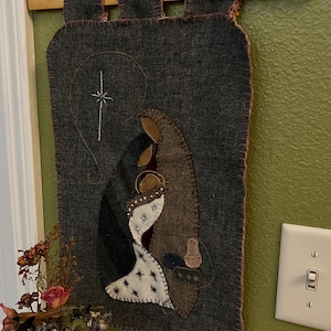 May include: A gray felt Christmas wall hanging with a quilted nativity scene. The hanging is attached to a wooden sign that reads "For unto us a child is born".