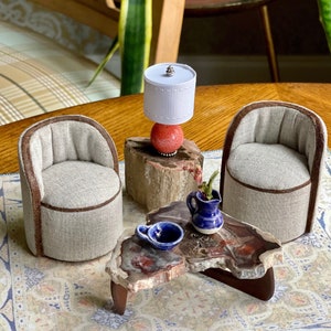 Könnte beinhalten: Ein Miniatur-Wohnzimmerset mit zwei beigen Sesseln, einem kleinen Holz-Couchtisch mit einer blauen Keramikvase und einer blauen Keramikschale sowie einer weißen Lampe mit rotem Sockel auf einem Holzstamm.