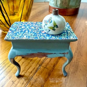 Könnte beinhalten: Ein kleiner Tisch im Antik-Stil mit einer verwitterten, tealblauen Oberfläche. Die Tischplatte zeigt ein blau-weißes Blumenmuster. Auf der Tischplatte steht eine kleine, weiße Vase mit Schmetterlingsillustrationen. Der Tisch hat geschwungene Beine.