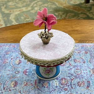 May include: A miniature pink flower sits on a white and pink cake stand with a blue and white base. The stand is decorated with a white lace trim and sits on a blue and purple patterned rug.