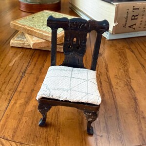 Puede incluir: Silla de madera en miniatura con acabado oscuro y cojín de asiento claro estampado. La silla tiene tallas ornamentales en el respaldo y las patas, mostrando un diseño clásico. Esta pequeña silla es un artículo decorativo, ideal para una casa de muñecas.