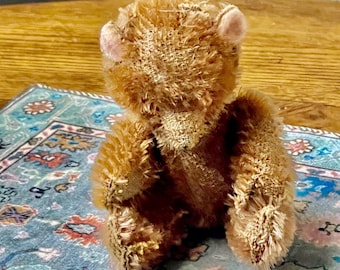 May include: A small, brown teddy bear with a worn, fuzzy texture. The bear is sitting on a blue and red patterned rug.