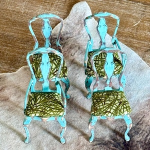 May include: Two miniature turquoise chairs with distressed paint, featuring curved legs and backs. The seats are upholstered with a green and white patterned fabric. The chairs are decorative and likely for a dollhouse or display.