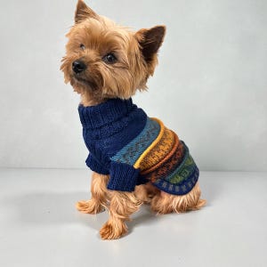 以下が含まれることがあります： 小さな犬が、ネイビーブルーとマルチカラーのセーターを着ています。セーターはリブ編みのタートルネックで、青、緑、黄色、オレンジの色合いの水平ストライプが特徴です。犬は茶色の毛並みで座っています。