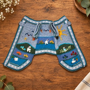 Peut inclure: Un vêtement en tricot bleu clair orné de paysages brodés avec des moutons blancs, un soleil jaune, des collines vertes et des fleurs colorées. La pièce présente une surface texturée avec des bordures décoratives et deux boucles à la taille.