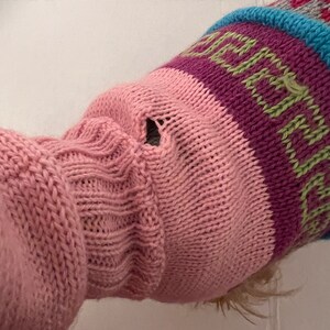May include: Close-up of a pink knitted sweater sleeve with a hole. The sleeve features a ribbed cuff and a patterned upper section with purple, green, and blue stripes. The sweater appears to be handmade.