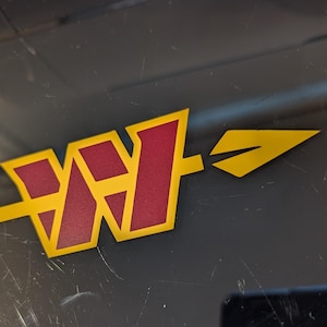 May include: Close-up of a logo featuring a stylized "WH" design in burgundy with a gold outline. A gold arrow extends to the right. The logo is set against a dark, possibly black, background with some scratches.