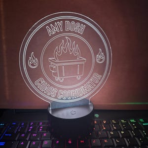May include: A clear acrylic LED lamp with the text "AMY DOSH CHAOS COORDINATOR" in a circular design. The design includes a dumpster with flames and is mounted on a black base. The lamp is placed on a laptop keyboard.