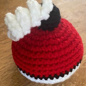 May include: A red, white, and black crocheted hat with a white pom-pom on top. The hat is shaped like a ball and has a black and white trim around the bottom.
