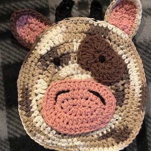 May include: A crocheted cow-shaped dishcloth with brown, white, and pink yarn. The cow has a brown nose, black eyes, and pink ears.