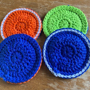 May include: Four crocheted coasters in different colors: orange, green, blue, and white. The coasters are round and have a white border.