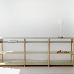 Op de afbeelding: Een lichtgroene en houten plank met drie planken. Een witte wereldbollamp staat op de bovenste plank. Een boek en een decoratief item staan op de planken. De plank staat tegen een witte muur.