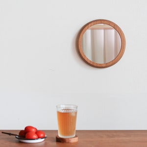 May include: A round wooden mirror with a natural wood finish is mounted on a white wall. Below, a glass of iced tea sits on a wooden coaster, and a small plate of red tomatoes rests on a wooden table.