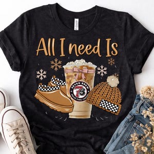 Puede incluir: Camiseta negra con el texto "All I Need Is" encima de una bebida de café, zapatos y un gorro. La taza de café tiene un lazo rosa y el logo de Seven Brew. El gorro y los zapatos tienen un patrón de cuadros blancos y negros.