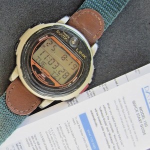May include: A Timex Data Link watch with a digital display showing the date and time. The watch has a silver-colored case, a brown leather strap, and a green fabric band. The watch is water-resistant up to 100 meters. A quick start guide is also visible.